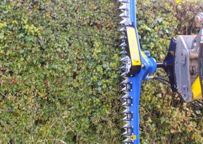 Image of the Slanetrac HC180 Mini Digger Finger Bar Hedge Cutter attachment cutting a hedge in a vertical position